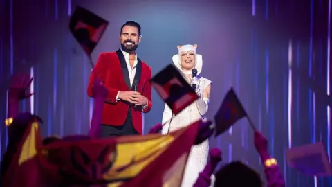 BBC Studios/Bad Wolf/James Pardon Rylan Clark, who has dark slicked-back hair, a dark beard and moustache and is stood on a stage, smiling at an audience of whom you can just see their arms in the air, waving flags. He is wearing a red velvet blazer and white shirt and is holding a microphone. Behind him is a person wearing a white dress and cape and animals ears, who is also holding a microphone.