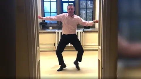 A man wearing a pink shirt and black trousers dances in a corridor, seen through an open doorway.