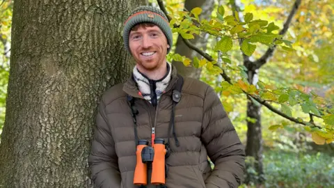 Jack Baddams Man standing in front a tree with bright orange binoculars. He is wearing a brown puffer jacket and a green beanie.