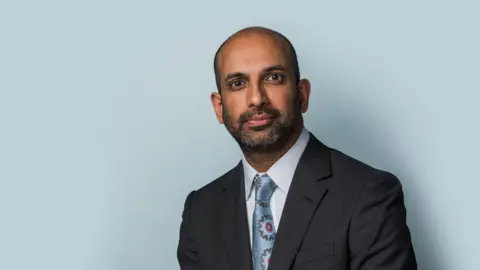 Rajiv Menon KC, who has a beard and brown eyes, standing in grey suit near blue wall.