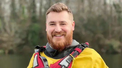 Marcus Fouracres Luke, a young man, wearing yellow and red wading equipment. He is smiling at the camera and has a ginger beard, brown hair and dark eyes.