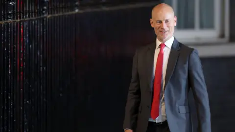 Getty Images A bald man in a dark suit, white shirt and red tie. 