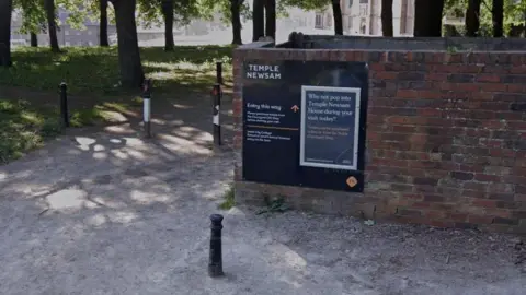 A sign on a brick wall indicating the way to Temple Newsam. Traffic bollards and trees can be seen in the background.