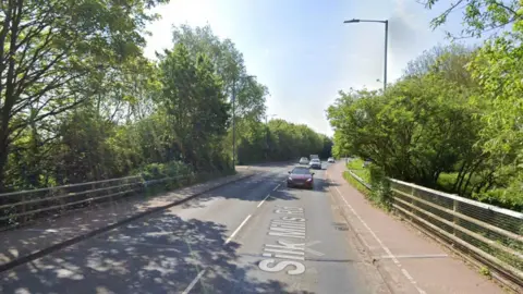 Google maps street view of Silk Mills Road in Taunton. There are railings and trees either side of the road and cars can be seen on the carriageway.