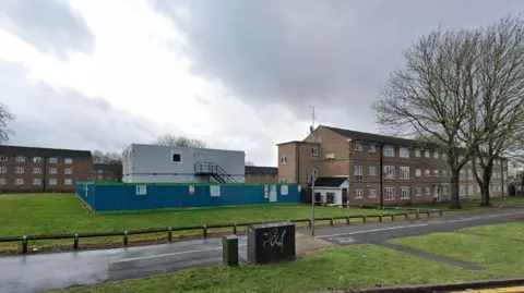 Google Maps Brown brick blocks of flats surround a green space, on which sits a grey temporary building, surrounded by blue fencing.