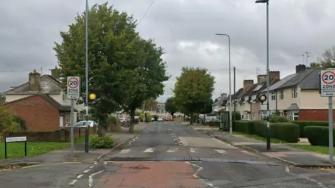 Image of a tree lined street with terraced housing on either side of the road. In the foreground can be seen a zebra crossing.