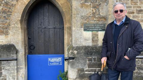 John Rigg next to a flood level sign and blue floodgate in a church doorway