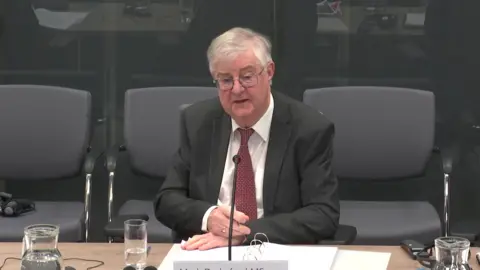 Senedd Cymru Mark Drakeford sat at a Senedd committee wearing a black suit, with a ring binder on the table in front of him and a glass of water.