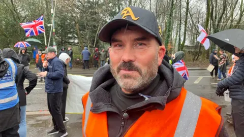 A man in a high-vis vest and a baseball cap, looking at the camera, close up. Several people are dispersed across the road behind him.