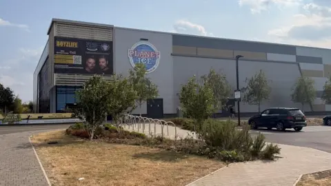 Picture of a building - with a sign on the wall which says "Planet Ice" a road runs in front of the building. 