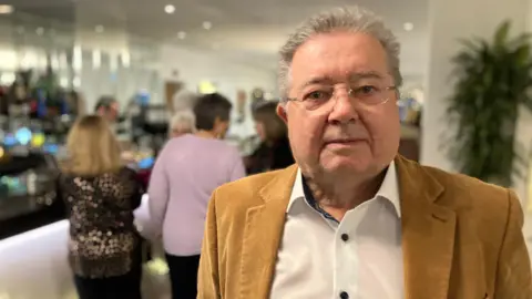 A man stands in the entrance foyer of a hotel. There is a reception desk and bar in the background, with customers walking through the foyer. 