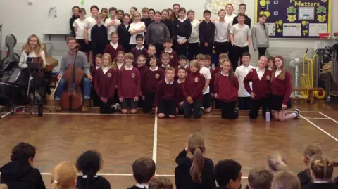 Mundella Primary School A large group of school children stand in rows alongside a group of musicians in a school hall. They are watched by more school children who are sitting in rows.