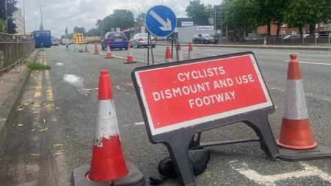 A sign asking cyclists to dismount along a coned-off section of the A56 Chester Road in Stretford