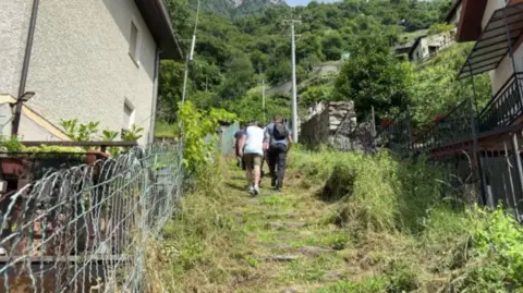 Phillip Norton/BBC Two men with some other people in front can be seen in action walking up steep rural steps covered in greenery to what can be seen to be several trees and a mountainous area up ahead. Either side of the trail are houses.