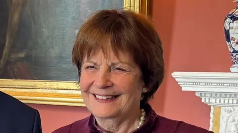 A head and shoulders image of Lady Clare Fitzroy, Countess of Euston, wearing a burgundy top and a pearl necklace. She is smiling and looking past the camera.