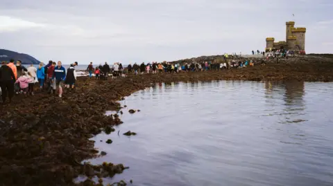 Hundreds of people walking out along a seaweed covered path to the Tower of Refuge, which is a small castle-like stone-built structure.