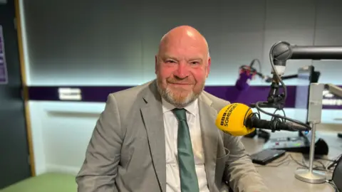 Bill Revans in a tie and suit jacket sitting behind a yellow BBC Radio Somerset microphone