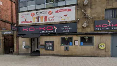 Street view image of the Ko-Ko Lounge, a stone building with two small square windows to the right of the entrance which is under a black canopy and a bright pink on black sign saying Ko-Ko Lounge