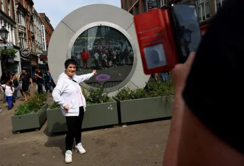 Hannah McKay/REUTERS  A woman poses in front of The Portal, a public technology sculpture that links with direct connection between Dublin, Ireland and the Flatiron district in Manhattan, New York City, in Dublin, Ireland, May 21, 2024.
