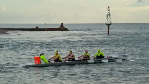 Five women sit in a coxed rowing boat in the sea. Four have oars while a fifth sits at the stern, captaining.