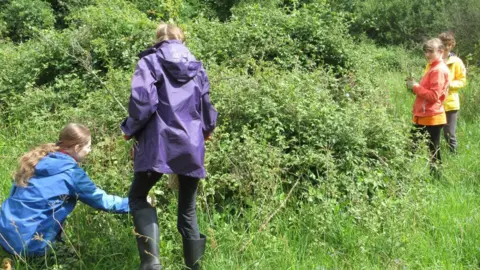 Norfolk Wildlife Trust Three girls dressed in bright jackets are supervised by an adult in wellies and a purple coat as they look and inspect overgrowth in a field.