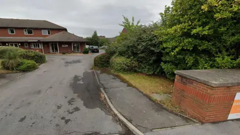 A view of Darnall Grange nursing home in Sheffield, with a long road leading up to the entrance of the two-storey building. A side and rear car park is visible in the distance. 