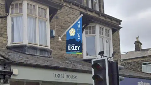 Row of shops on the ground floor and on the first floor a short flag pole juts out and from it hangs a flag in shades of blue showing the Yorkshire rose, the words Na Then and promotion for the town of Ilkley