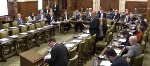 UK PARLIAMENT Room with wooden panels and green carpet with wooden tables and chairs, several people sat around a rounded wooden table, bald man in a suit stood up speaking