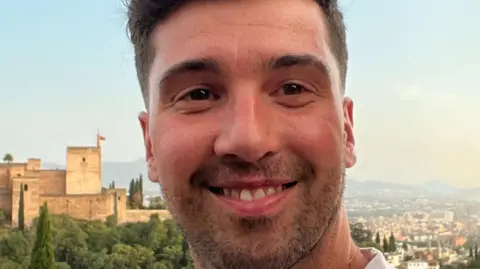 Number Nine PR Ben smiles at the camera while standing on a hill above a castle and a town below him. He has short brown hair and a stubbly brown beard.