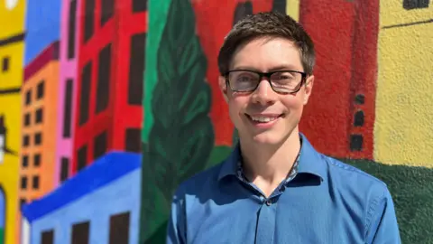 Sue Dougan/BBC Head and shoulders image of Sam Wakeford standing in front of a colourful wall. He has short dark hair, is wearing glasses and a blue shirt.