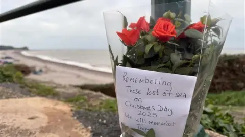 A floral tribute tied to a pole. On the distance there is a beach. The flowers are read and there is a note in the flowers with the words 'Remembering those lost at sea on Christmas Day 2025. We will remember them.'