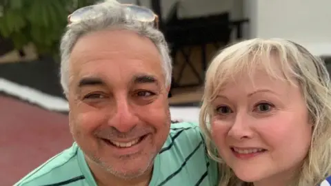 Rachel Hyman A man on the left wearing a green t-shirt. He has grey hair and his left arm around a middle-aged blonde woman. Both are smiling.