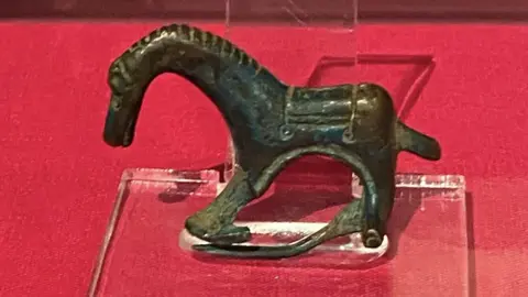 A small copper-alloy horse figurine which has a long curved neck is propped up on a clear stand on a red table.