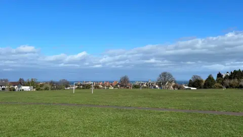 Wide open grassy field at Joppa Quarry Park with two white football goals, a paved path in the foreground, houses and trees in the distance, and a blue sky with scattered clouds above.