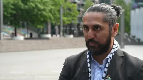 Ihtesham Afzal in a blue and white stripe shirt, a black blazer, a black and white scarf and a Palestinian flag pin on his lapel.