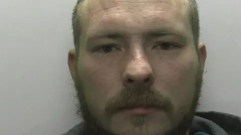 Police mugshot of Luke Cowling who has a beard and is wearing a grey hoodie