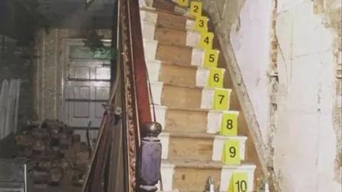 PA Media The stairs in Bleak House. There are wooden stairs and a banister to the left. There are crime scene numbers on each step. In the background is a door and rubble on the floor