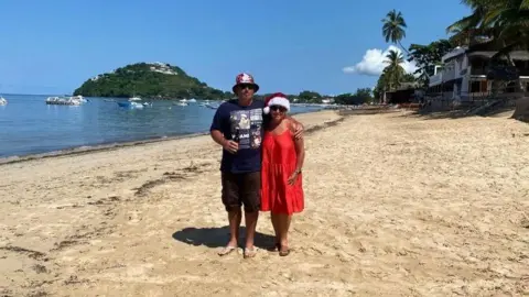 Victoria Reitze The couple are on a beach in Madagascar, wearing summer clothes and Victoria is wearing a Christmas hat. There are small boats and palm trees in the background.