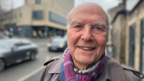Elizabeth Baines/BBC A man with white hair and eyebrows wearing a grey jumper and coat with purple scarf. 
