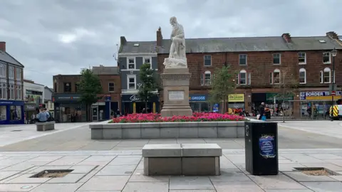 Statue of Robert Burns surrounded by flower beds in Dumfries. Behind the statue is a row of shops, including Greggs and Border Travel