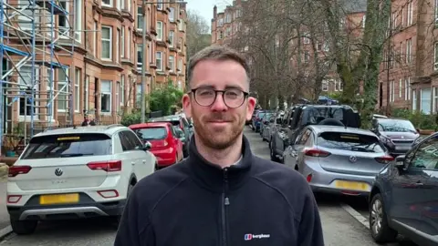 david kelly wearing glasses and a dark outdoor jacket stands in the foreground of a residential street lined with parked cars and red-brick buildings. Scaffolding is visible on one building to the left, and trees without leaves line the street in the background.