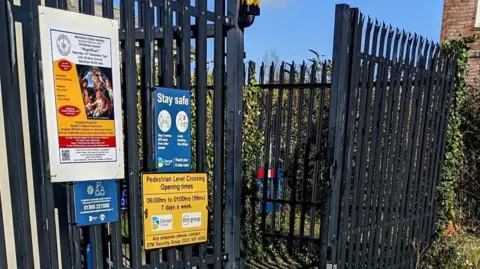 Jaggery Tall metal fencing and gates at the entrance to the railway level crossing at Wareham. Signs on the black metal indicate the crossing is open from 6am to 1am, seven days a week.