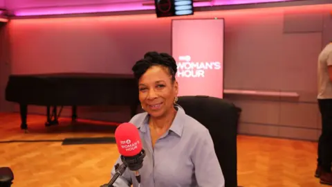 Kimberle Crenshaw has dark hair and is wearing a light purple shirt. She is sat infant of a red radio microphone.