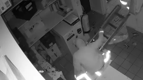 Black-and-white CCTV footage shows Matthew Pickering trying to stand up after falling from the ceiling. A ladder and a high visibility jacket are on the floor. Pickering is wearing a sweatshirt with a hood covering his head.