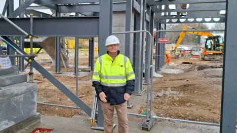 Man in white hard hat and high-visibility jacket stands on muddy ground at a construction site, facing a steel framework structure with beams and columns; excavator and building materials are visible, with a "Do Not Enter" sign on a staircase and temporary fencing surrounding the area.