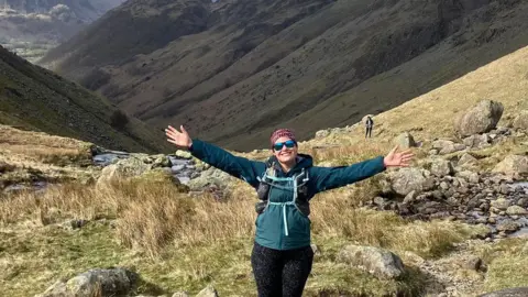 Sophie Hatfield Runner Sophie Hatfield stands with her arms wide open in the Northern fells. She has brown hair and is wearing sunglasses, a pink buff headband, a green waterproof jacket and black leggings. 