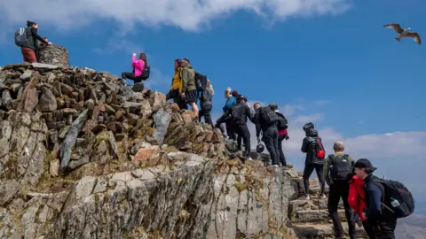 People wearing coats and backpacks queue to reach the top of a mountain, against the backdrop of a blue sky.