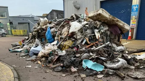 A large pile of rubbish that has been fly-tipped on an industrial estate outside a garage. There is bedding, mattresses as well as wood in the dump.