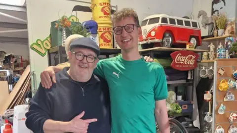 Two men in glasses, each with an arm around the shoulder of the other. They are both smiling, the older man is wearing a flat cap