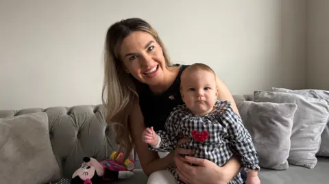 Chloe Heath in a black top with long blonde hair. She is sat on a grey sofa with grey cushions and is holding a baby, who is wearing a chequered dress with a red heart on the front.